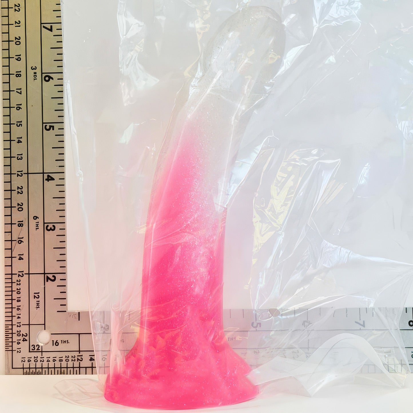 A pearl sparkle rose silicone dildo wrapped in clear plastic, shown next to a metal ruler for scale on a white background.