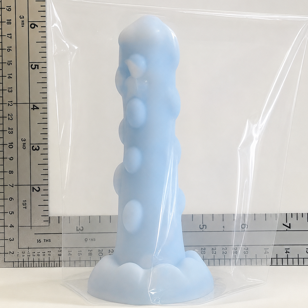A light blue silicone adult novelty product, likely a suction-base dildo, sealed inside a clear plastic bag with a ruler visible behind it for scale.