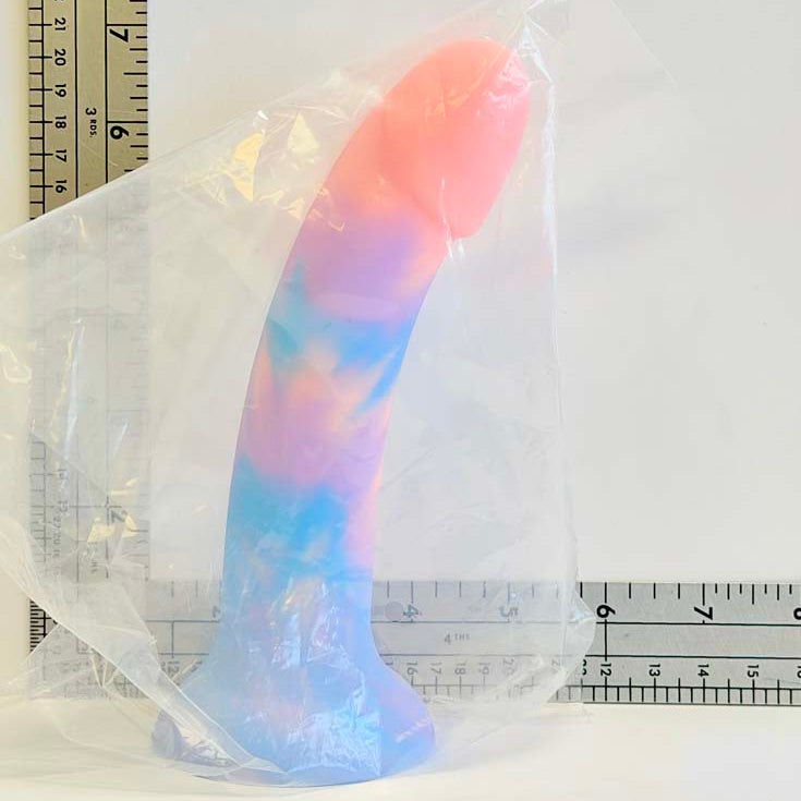 A curved silicone dildo wrapped in clear plastic packaging, shown with metal rulers for scale against a plain white background.
