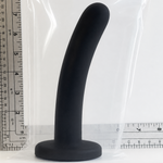 A black curved silicone plug with a wide circular base, shown standing upright next to a clear plastic bag and a ruler for scale on a white background.