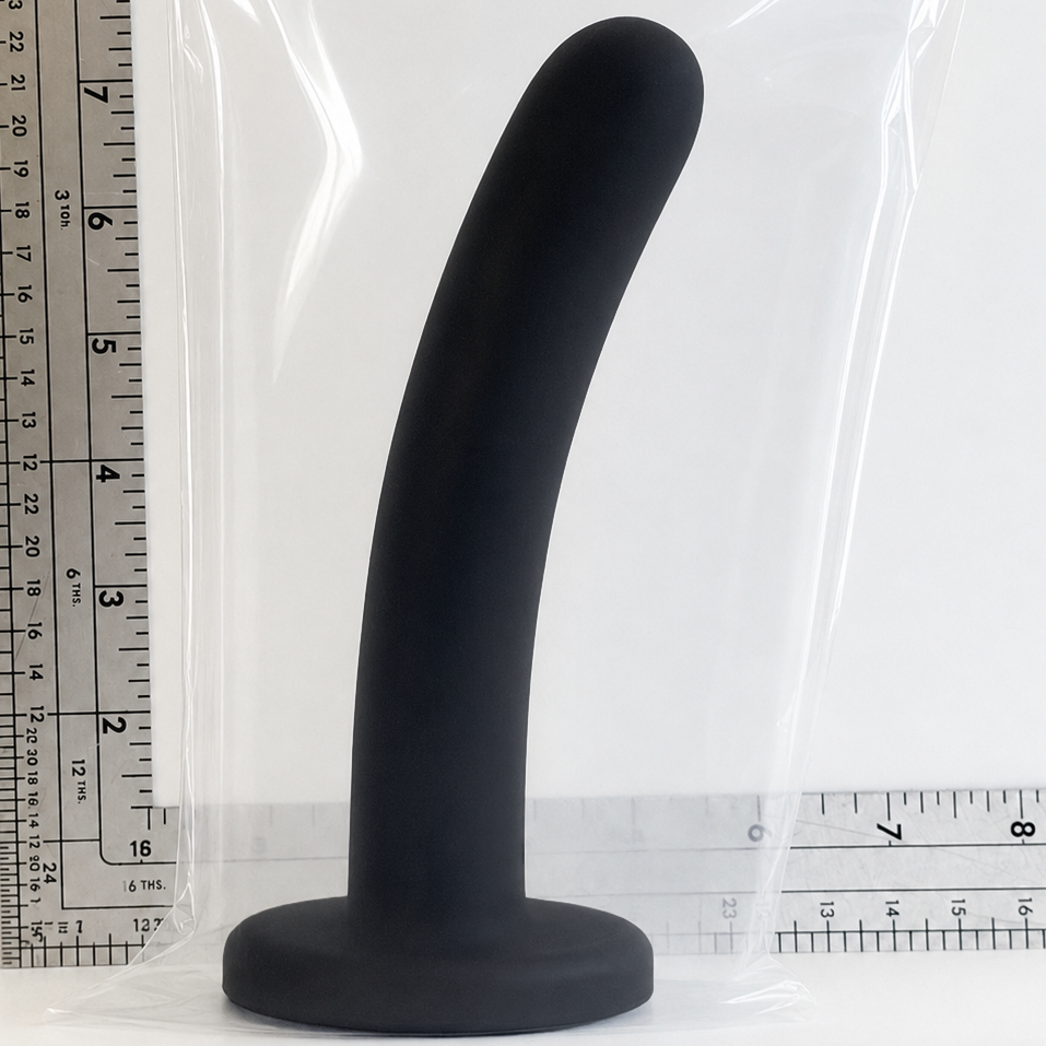 A black curved silicone plug with a wide circular base, shown standing upright next to a clear plastic bag and a ruler for scale on a white background.
